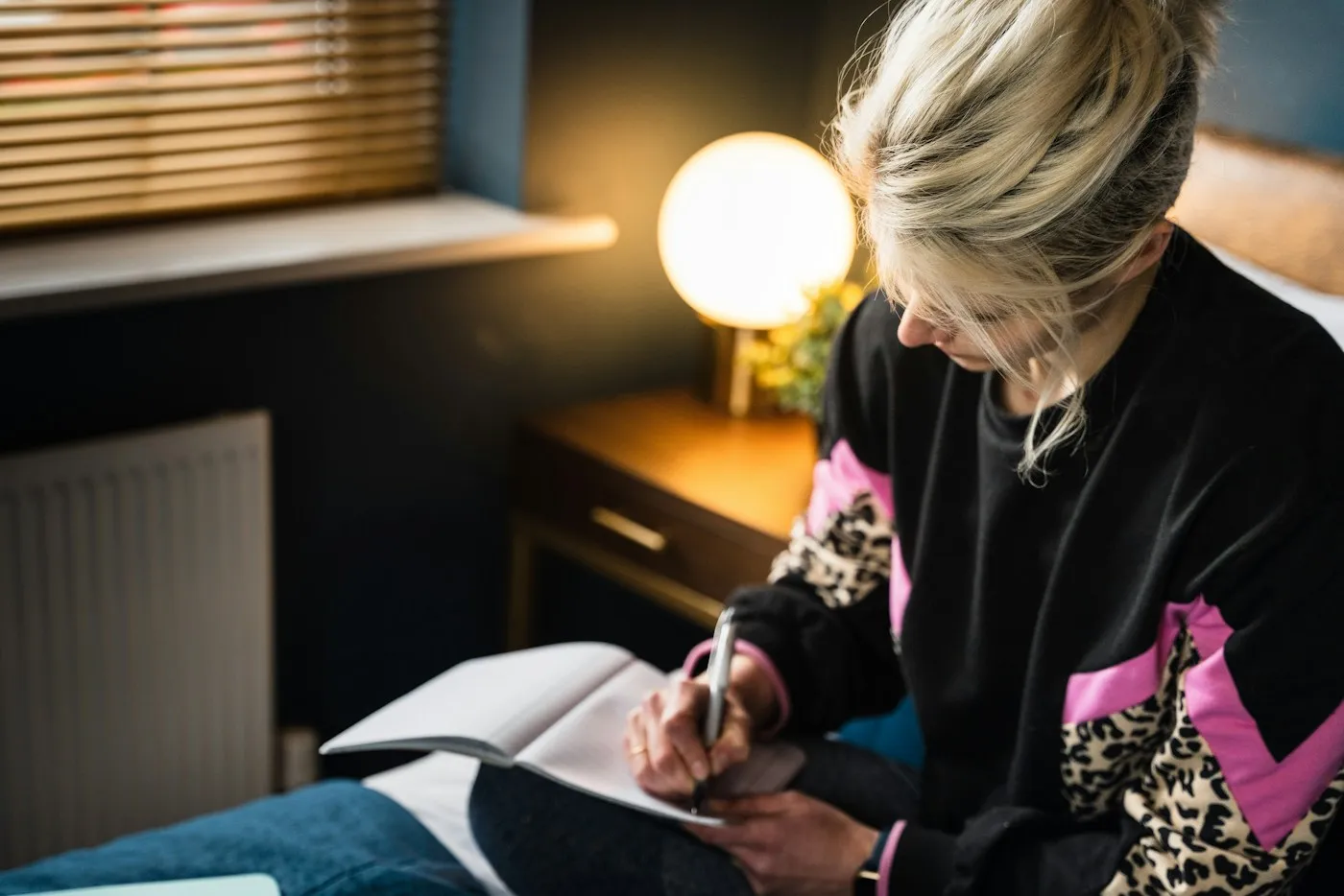 Woman writing in a notebook on her bed by the glow of a bedside lamp as part of a calming bedtime routine