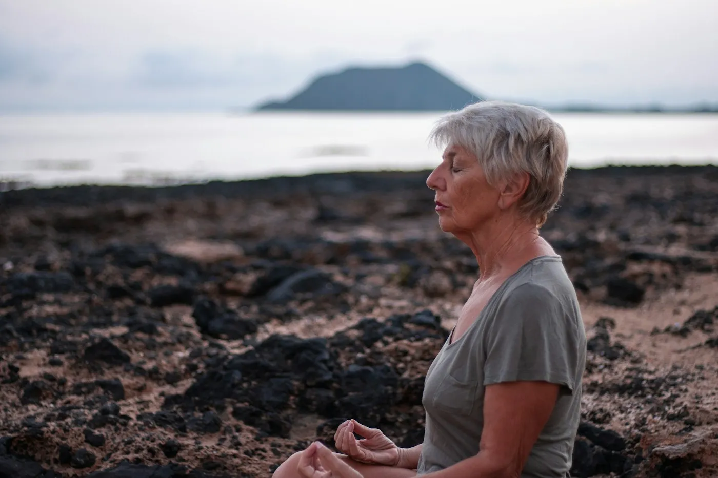 Older woman with short gray hair meditating calmly on a rocky shore at dusk with her eyes closed
