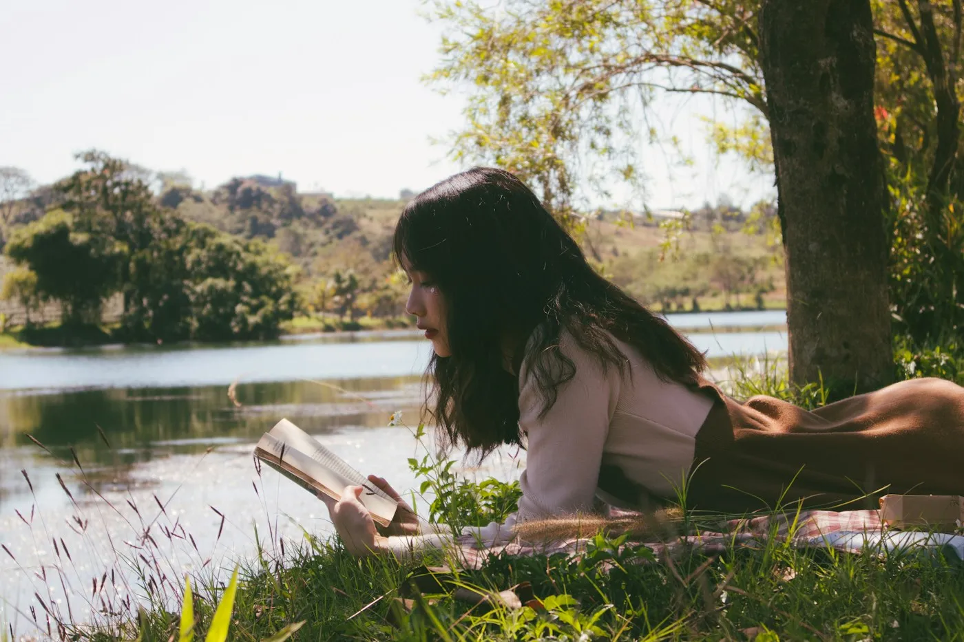 Woman lying on the grass by a calm lake reading a paper book, enjoying a digital detox