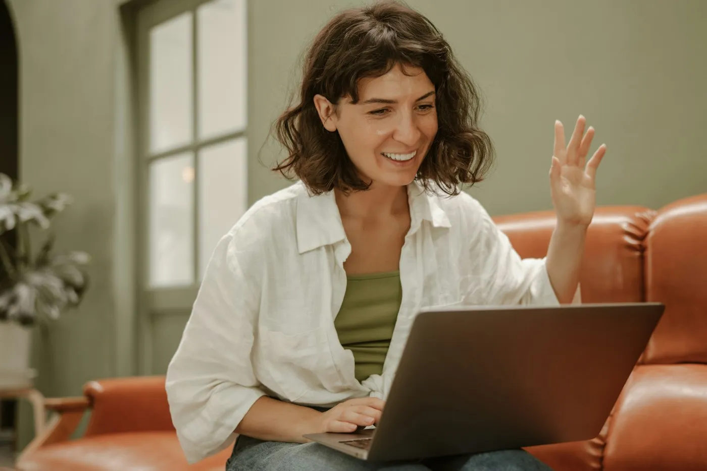 Smiling woman on a sofa waving at her laptop during a first video call with a therapist