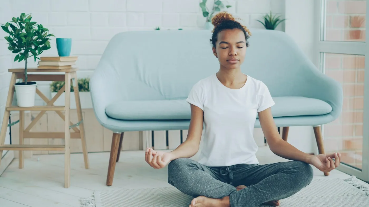 Young woman meditating cross-legged on a rug at home with her eyes closed and hands resting on her knees