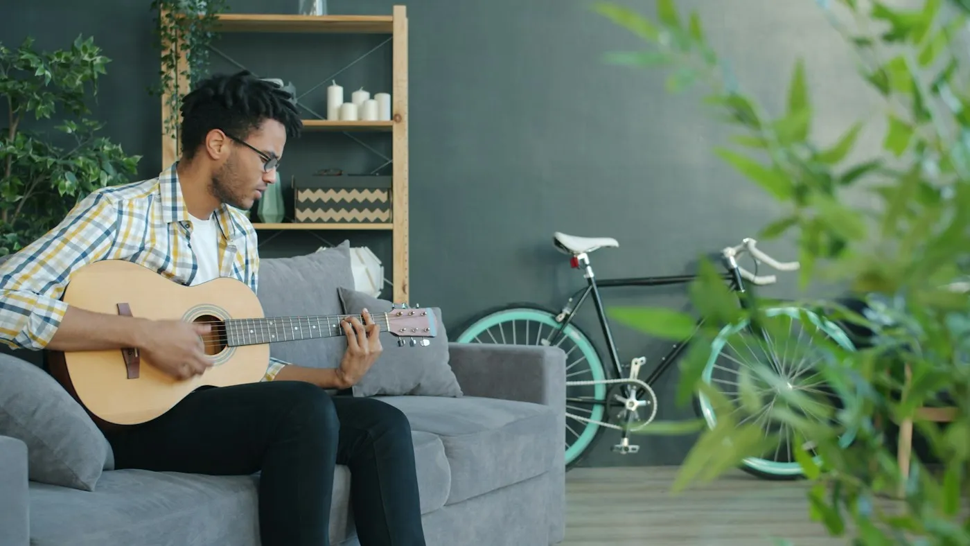 Man in a plaid shirt relaxing on a gray sofa at home while playing an acoustic guitar after work