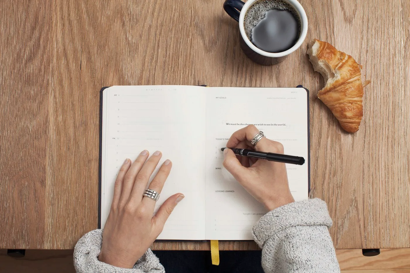 Hands writing in an open gratitude journal on a wooden table with coffee and a croissant