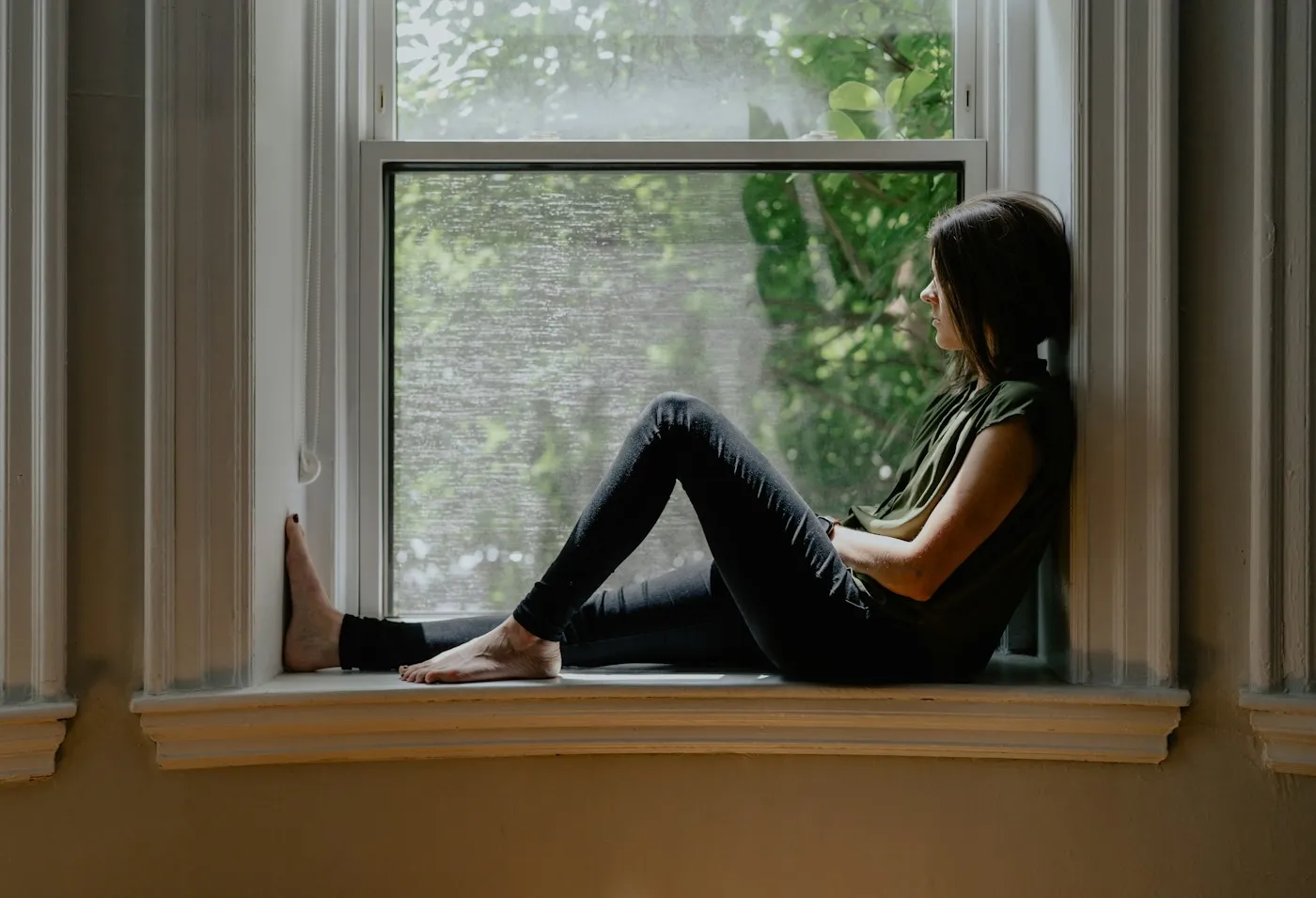 Woman sitting quietly on a sunny window seat, looking outside and reflecting