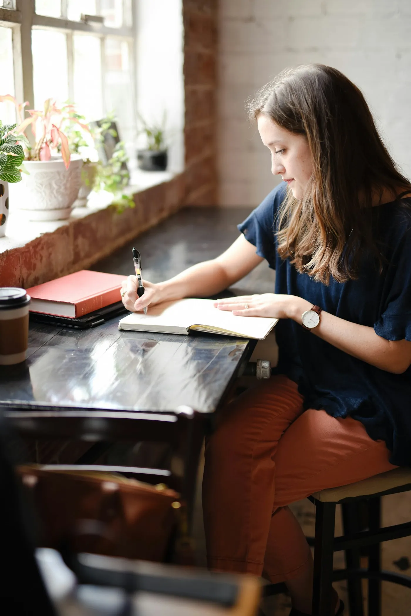 Young woman writing in a notebook at a sunny cafe table by a window with plants