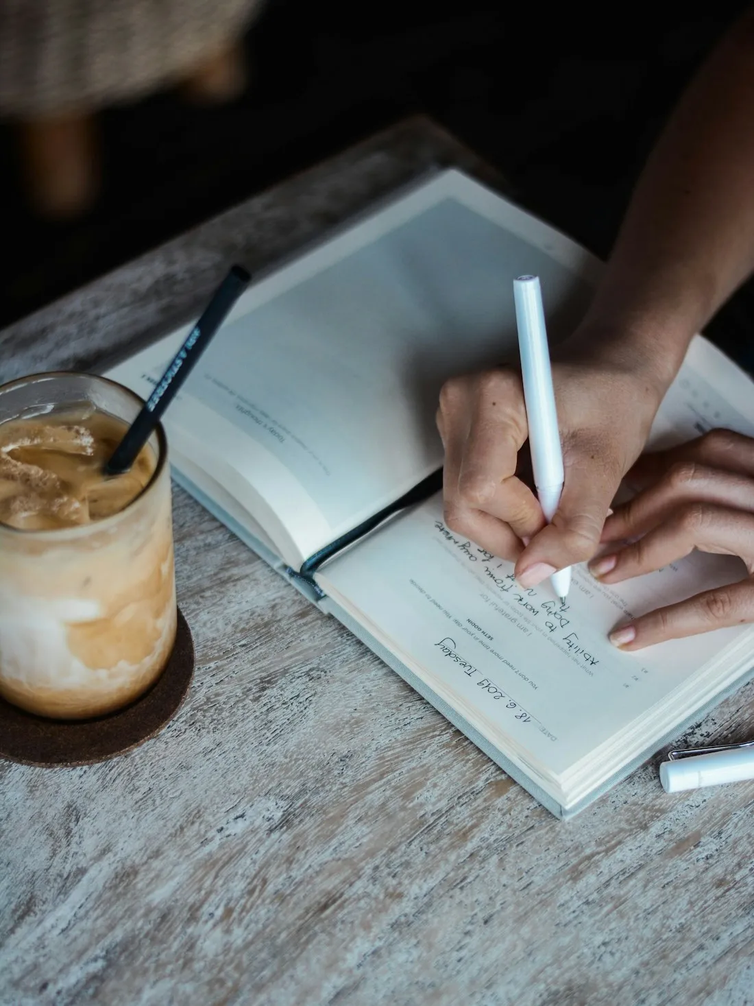 Hand writing a dated journal entry in an open notebook beside an iced coffee