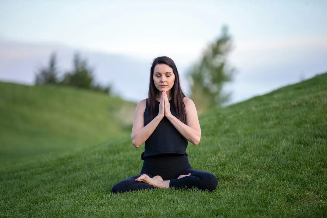 Woman meditating cross-legged on a grassy hill with her eyes closed and palms pressed together