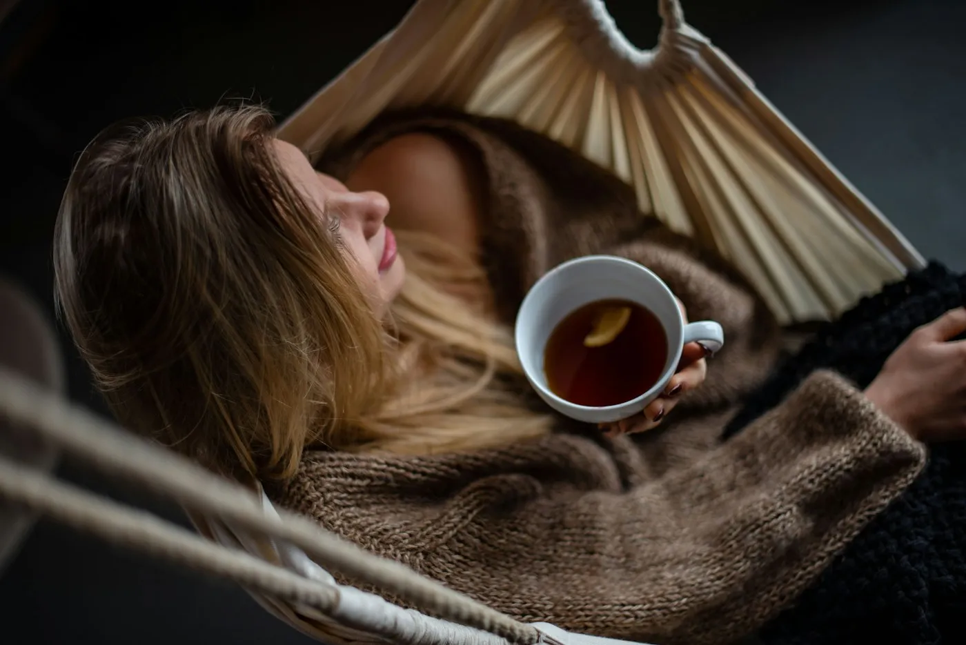 Woman resting in a cozy hammock with her eyes closed, holding a cup of tea with lemon