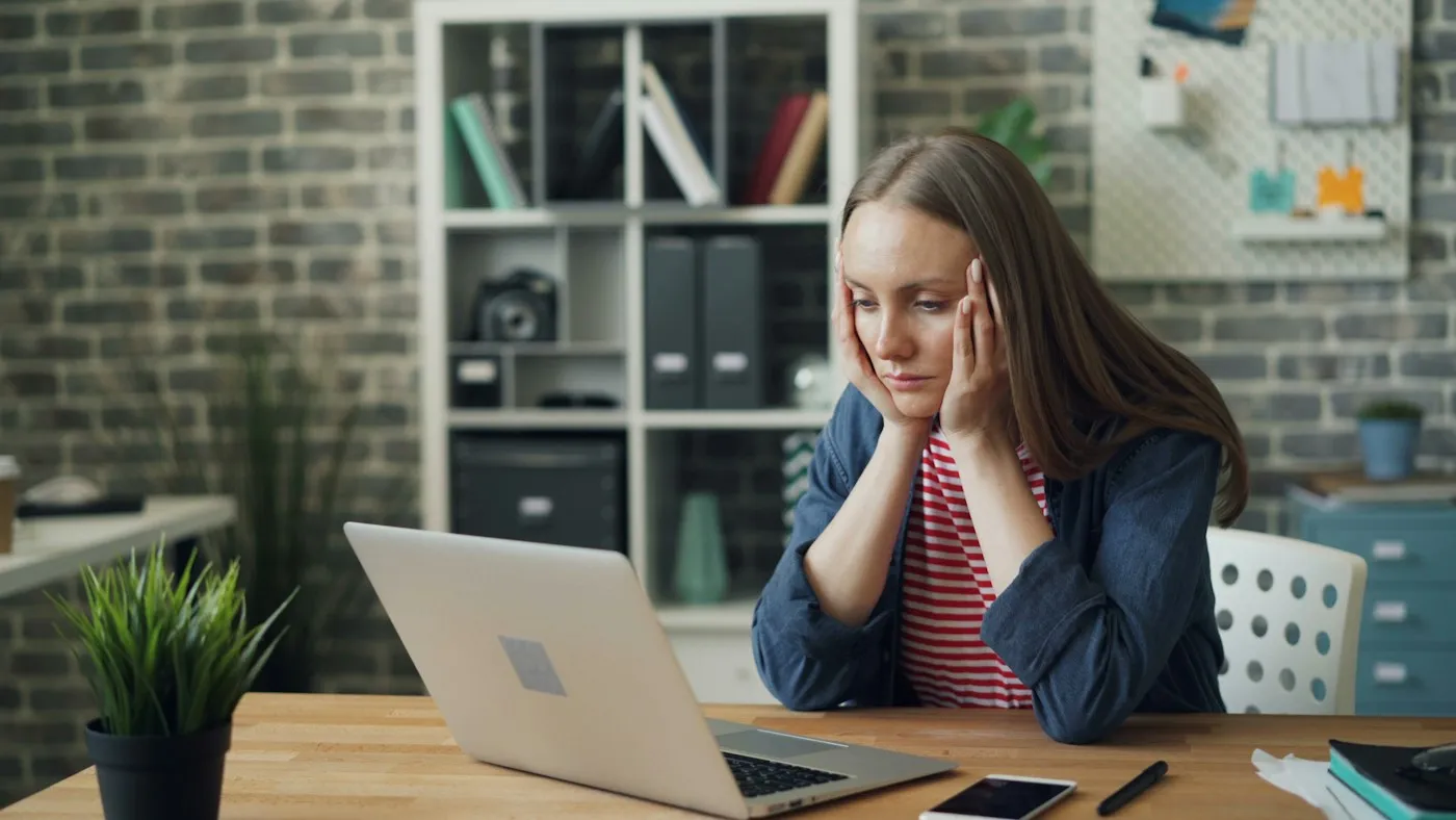 Tired young woman resting her head in her hands at a laptop in a bright office, showing common signs of burnout