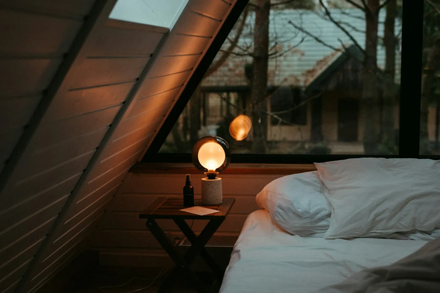 Calm attic bedroom at dusk with a warm bedside lamp and fresh white bedding, set up for good sleep hygiene