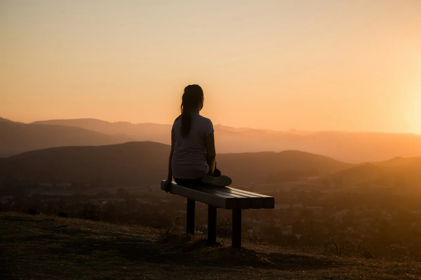 Person sitting alone on a bench on a hilltop at sunset, looking out over the valley in quiet reflection