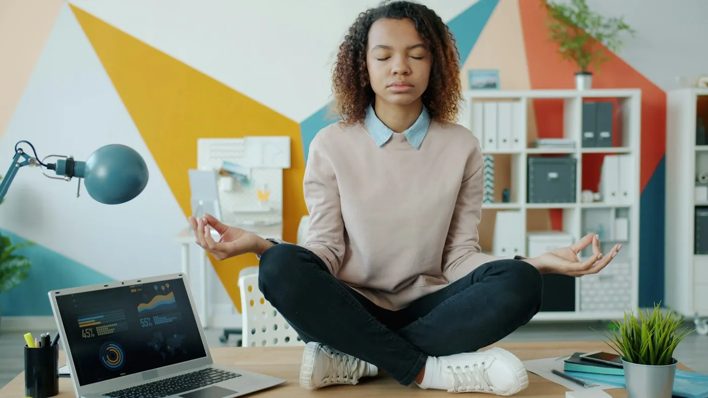 Young woman meditating with closed eyes on her desk beside a laptop, taking a calm break at work