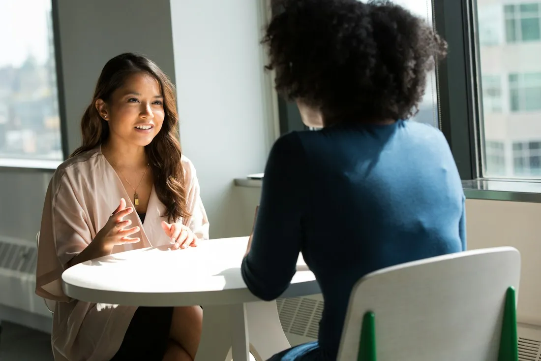 Two women talking at a small round table by a sunny window during a supportive conversation