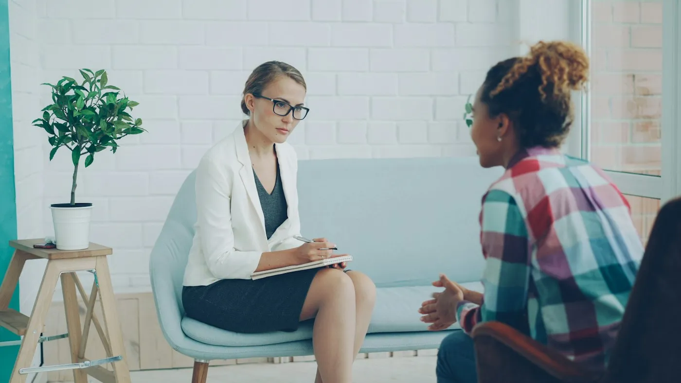 Therapist in glasses taking notes while listening to a young woman in a bright counseling room